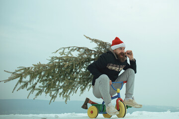 Hipster carrying Christmas tree on white snow. Excited crazy funny cyclist in santa hat celebration Christmas and new year. Man riding bicycle tricycle on snowy landscape