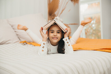 Portrait of a smiling happy little girl holding a book on her head and lying on the bed.