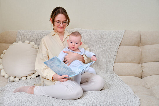 Happy woman mother with infant baby reading book on home sofa in living room