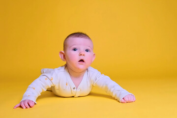 Infant baby boy looks in surprise while lying on his tummy, studio yellow background. Four month old child close up
