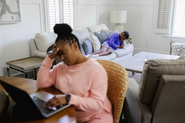 Woman using laptop at table in living room, teenage daughter lying on sofa in background
