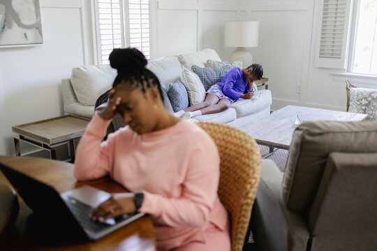Woman using laptop at table in living room, teenage daughter lying on sofa in background