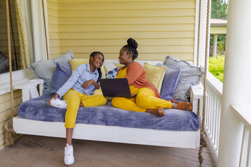 Smiling mother and teenage daughter sitting on porch swing with laptop