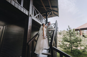 Beautiful, stylish newlyweds are standing on a wooden staircase in a large house. Wedding portrait, photograph of a young groom in a gray suit and a cute bride in a white dress.
