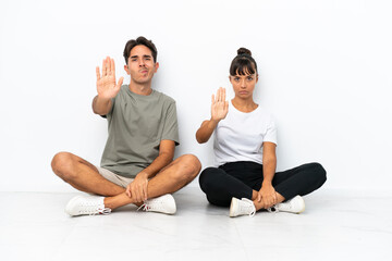 Fototapeta na wymiar Young mixed race couple sitting on the floor isolated on white background making stop gesture denying a situation that thinks wrong