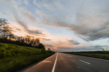 A broken car stands on the side of the road on a wet asphalt road after rain against the backdrop of a sunset and a blue sky with clouds. Beautiful landscape. Evening photography.