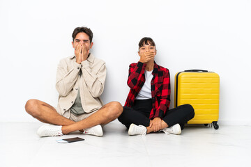 Young mixed race travelers couple sitting on the floor isolated on white background smiling a lot while covering mouth