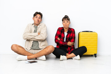 Young mixed race travelers couple sitting on the floor isolated on white background making doubts gesture while lifting the shoulders