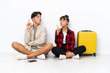 Young mixed race travelers couple sitting on the floor isolated on white background with fingers crossing and wishing the best