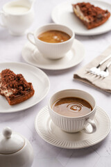 sweet breakfast - two white porcelain cups with coffee drink cappuccino with milk on a white saucer, piece of brown cake