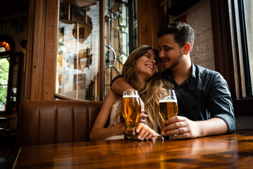 attractive young man and woman couple in love sitting down at pub indoors drinking draft beer and...
