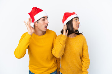 Young mixed race couple celebrating Christmas isolated on white background listening to something by putting hand on the ear