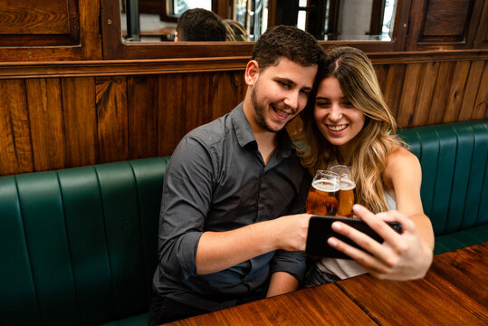 cute and young man and woman friends sitting down at indoor pub having fun, taking selfie and drinking draft beer - Powered by Adobe