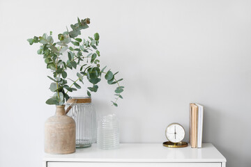 Vases with eucalyptus branches, alarm clock and books on shelf near light wall - Powered by Adobe