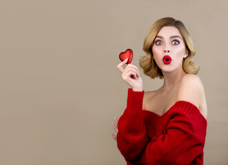 Young woman looks surprised to the camera and holds a red heart in her hands. Model with perfect young skin and professional makeup with red lips poses with red heart.  Christmas and New Year concept.