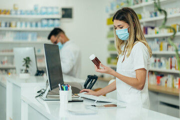 Pharmacist with protective mask on her face holding drugs and using computer