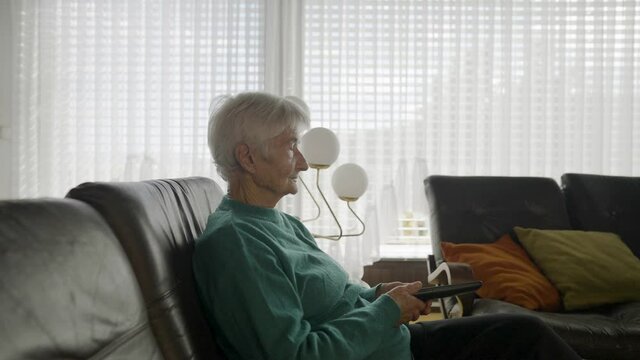 Slow motion - Zooming in on a grandma, in a crewneck and black pants, holding a TV remote, and looking at the television, with a window wall in the background, covered with shades, in daylight.