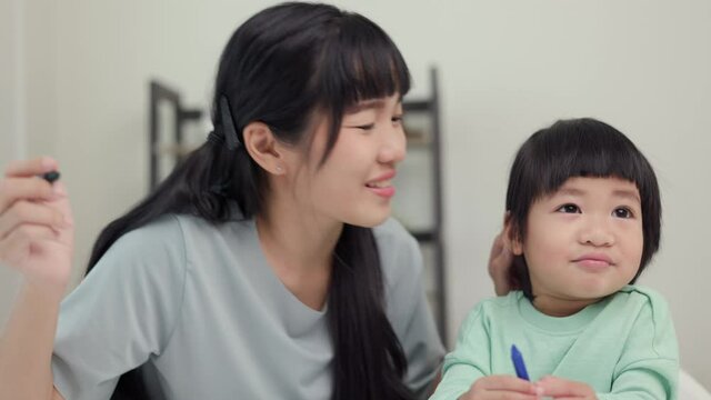 Mom kissing son and teaching how to painting with crayon color on book or doing homework. Happy asian boy painting with crayon with his mother in living room at home. Family having fun concept.