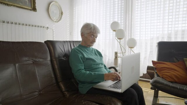 Mid shot - An older woman sitting in her living room on a leather sofa, holding a computer and typing on it