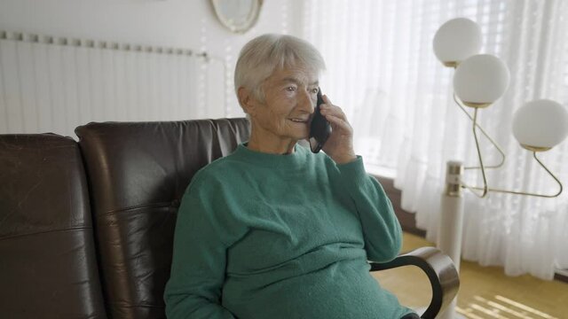 Medium shot - A grandma, in a blue crewneck, sitting on a leather sofa, in a living room with a big window with shades, holding a phone and talking with someone on it, with lights on her left side, on