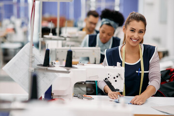 Young happy seamstress works at production line in factory and looks at camera. - obrazy, fototapety, plakaty