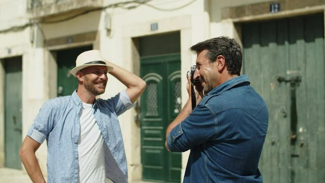 Medium shot of happy gay taking picture of lover on camera. Handsome homosexual man in hat posing for camera, smiling, enjoying time spending with boyfriend. LGBT, photoshoot concept