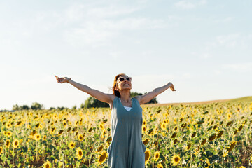 Naklejka na ściany i meble Middle-aged young woman wearing sunglasses, breathing fresh air with open arms in sunflower field