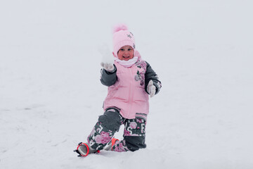 Baby for a walk in winter. Little girl in warm ski suit and snowmobiles on boots play in snow. 