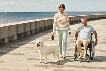 Portrait of adult couple with man in wheelchair walking dog outdoors by river, copy space - Powered by Adobe
