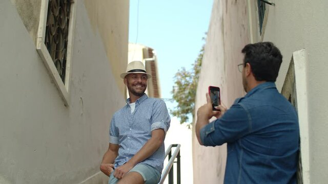 Medium shot of handsome gay taking picture of boyfriend outdoor. Smiling homosexual man standing in narrow street of old Spain village on summer day and posing for camera. LGBT, photoshoot concept
