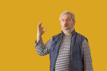 A man with a gray beard shows sign "OK". On a yellow isolated background.