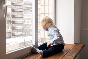 Adorable little blond kid boy sitting on the windowsill. A child looks out the window.