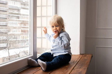 Child looks out the window. Adorable little blond kid boy sitting on the windowsill.