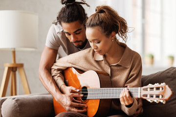 Smiling mixed race woman on sofa playing guitar for boyfriend while relaxing on weekend at home