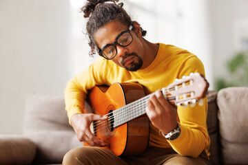 Young joyful african american man playing acoustic guitar at home, sitting on sofa in living room - obrazy, fototapety, plakaty