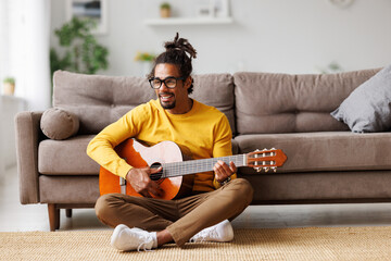 Young joyful african american man playing acoustic guitar at home, sitting on floor in living room
