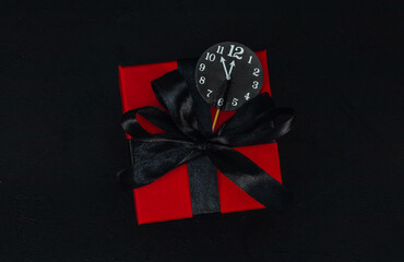 One small red gift box with a black ribbon and a paper clock in the middle on a black background