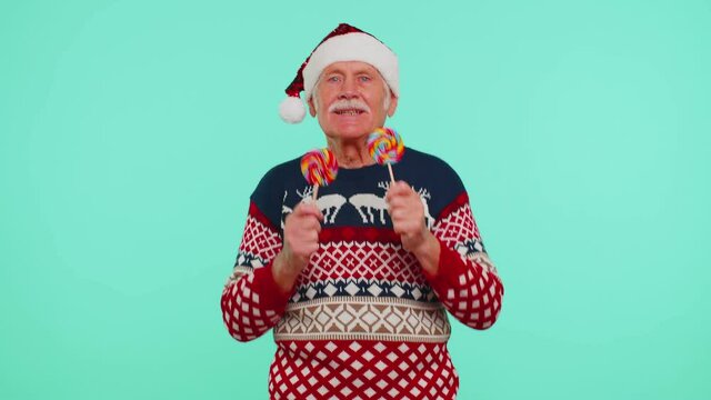 Senior grandfather old man in Christmas sweater, hat holding candy striped lollipops, fooling around, dancing, making silly faces isolated on blue background. Happy New Yearcelebration merry holidays