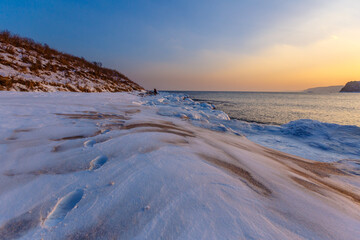 Colorful sunset on the snowy coast of the Sea of Japan. The Sonce goes over the snowy hills against the backdrop of the calm sea. Sikhote-Alin Biosphere Reserve in the Primorsky Territory.