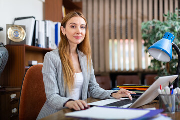Young female secretary in blouse sitting at desk and working on laptop. - obrazy, fototapety, plakaty