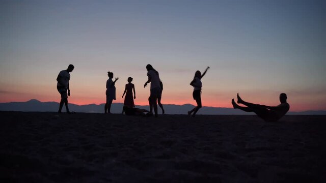 Cheerful guy showing sport tricks at the sea beach, crowd of friends dancing barefoot on the sand and enjoying amazing sea sunset. Silhouettes of happy dancing people at the beach party
