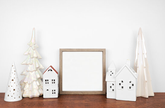 Christmas mock up with wood frame and white tree and house decor. Square frame on a wooden shelf against a white wall. Copy space.