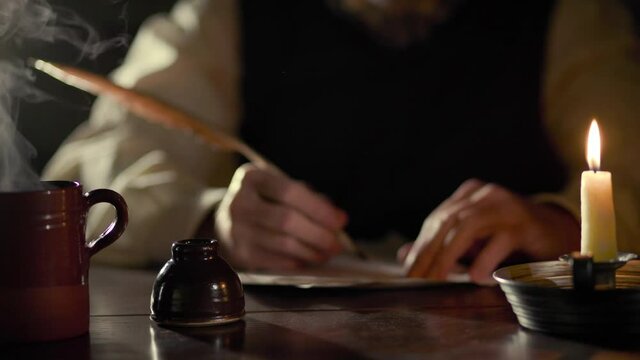 A scene from the 18th century of a man writing with a quill pen with a candle and a hot beverage sitting on his desk.