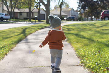 Toddler walking down the sidewalk in knitted sweater and hat in the fall; suburban neighborhood in...