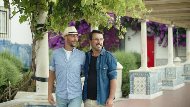 Gay couple walking in public garden with blooming trees. Front view of happy homosexual male lovers hugging each others waist, talking, strolling together during romantic date. LGBT, travel concept