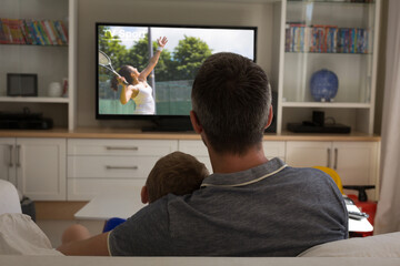 Rear view of father and son sitting at home together watching tennis match on tv