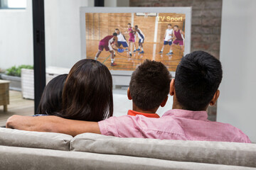 Rear view of family sitting at home together watching basketball match on tv