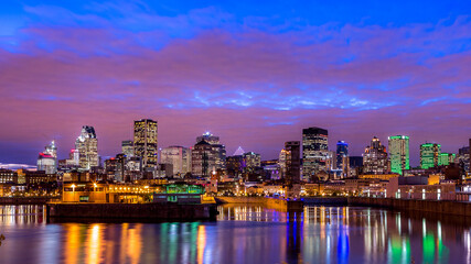 Fototapeta na wymiar Photo de la ville de Montréal prise au début de la nuit. Le ciel est d'un bleu intense, les nuages rosés et les lumières de la ville se reflètent sur le fleuve Saint-Laurent.