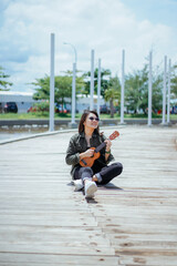 Fototapeta premium Playing Ukulele of Young Beautiful Asian Woman Wearing Jacket And Black Jeans Posing Outdoors