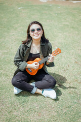 Playing Ukulele of Young Beautiful Asian Woman Wearing Jacket And Black Jeans Posing Outdoors